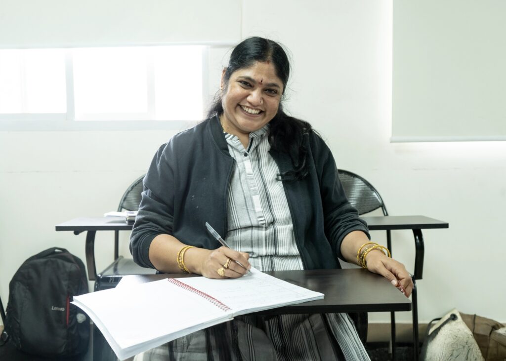 Sai Shree Suresh, a middle-aged woman with long black hair, wearing a striped dress and dark jacket, smiling while sitting at a classroom desk and writing in a notebook.