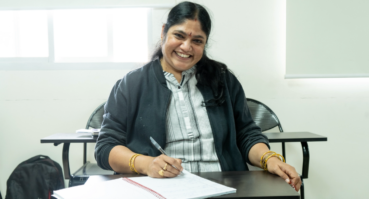 Sai Shree Suresh, a middle-aged woman with long black hair, wearing a striped dress and dark jacket, smiling while sitting at a classroom desk and writing in a notebook.