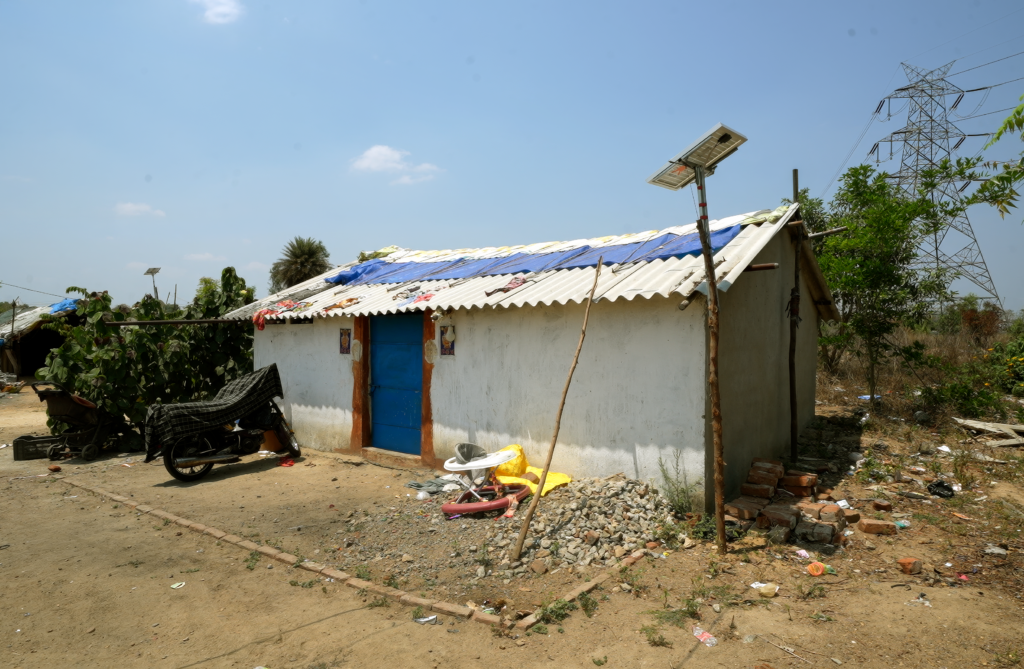 A small single-story house with white walls and a corrugated roof patched with blue tarpaulin. A solar streetlight stands beside it.