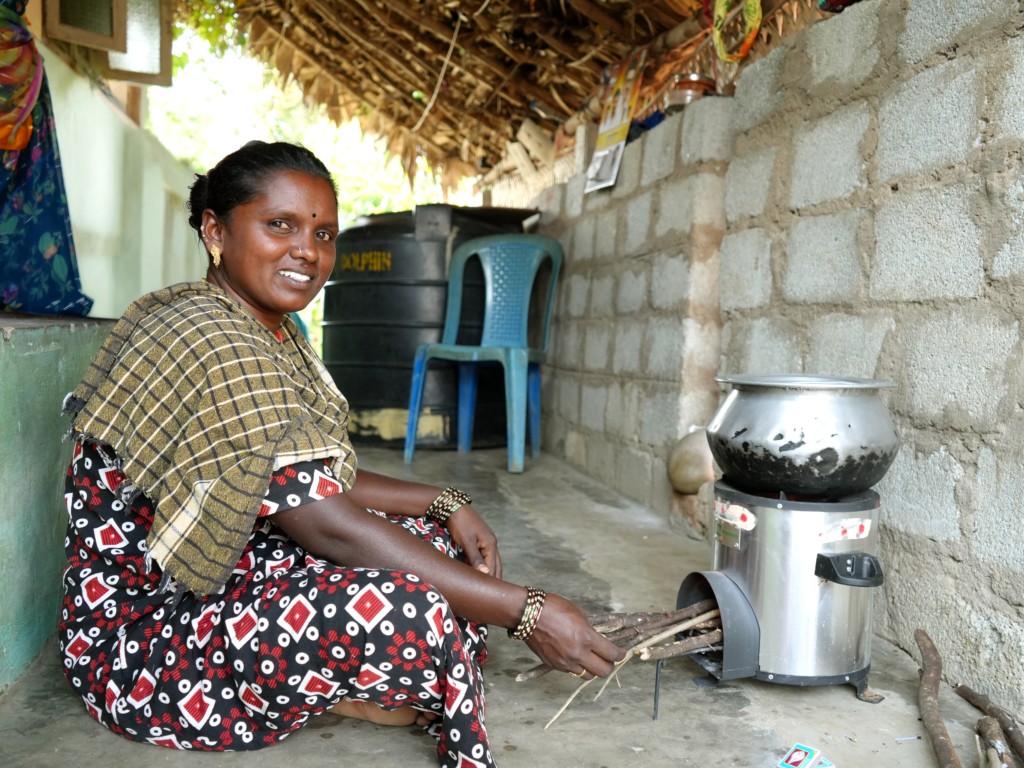 A woman sits on the ground in front of an improved cookstove with a pot on top, feeding wood sticks into the stove’s fuel opening.