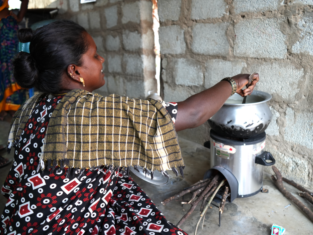 A woman sits on the ground in front of an improved cookstove stirring food in a pot.