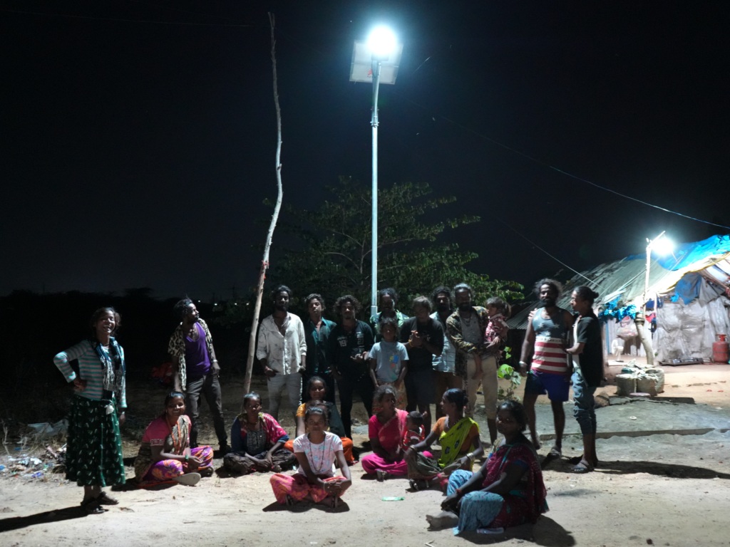 A group of men, women, and children sitting and standing under a solar streetlight at night