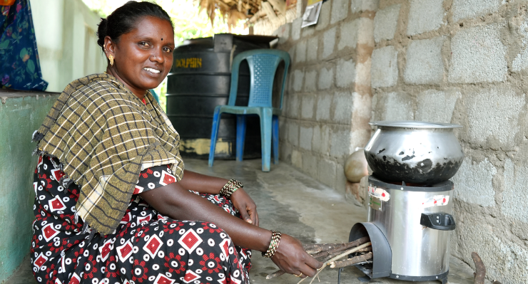 A woman sits on the ground in front of an improved cookstove with a pot on top, feeding wood sticks into the stove’s fuel opening.
