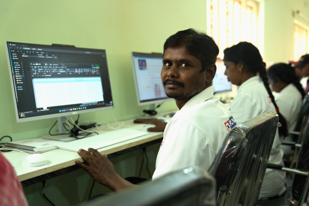 Venkateshappa sits at a computer in a training room, turning to look at the camera while others work at desktop screens beside him. He wears a white polo t-shirt.