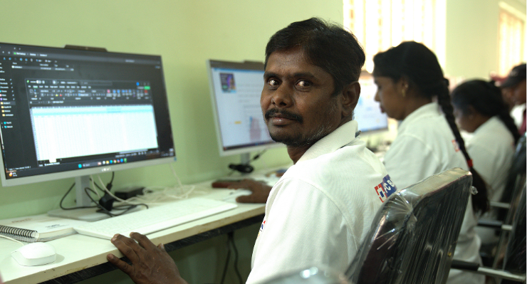 Venkateshappa sits at a computer in a training room, turning to look at the camera while others work at desktop screens beside him. He wears a white polo t-shirt.