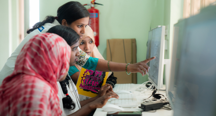 A woman is pointing at a computer screen explaining something to other two young women